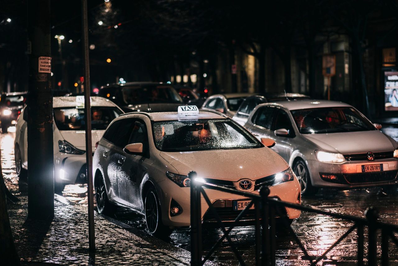 Home Busy city street at night with cars and taxis on a wet road, reflecting lights.